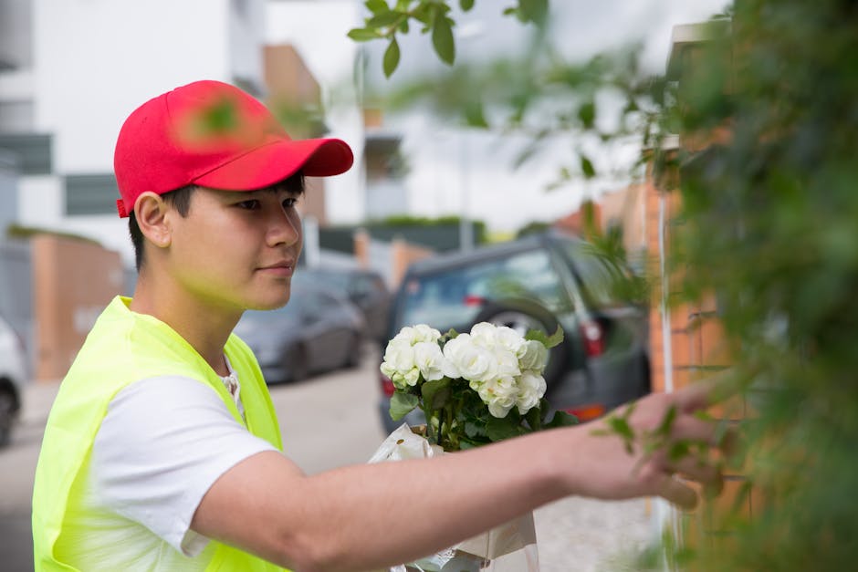 A professional florist, wearing a red cap and a high-visibility yellow vest over a white shirt, is delivering a floral gift outdoors on a residential street. He holds a bouquet of fresh white hydrange