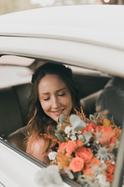 A young woman with long, wavy brown hair smiles softly as she holds a floral bouquet inside a car. The bouquet features a vibrant mix of peach and coral roses, white daisies, and greenery, all arrange