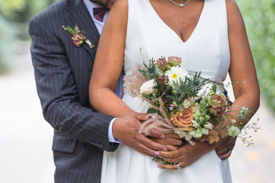 A bride and groom stand outdoors on a natural, blurred background, with the groom dressed in a dark pinstripe suit holding the bride, who wears a white gown with a V-neckline. The bride holds a large,
