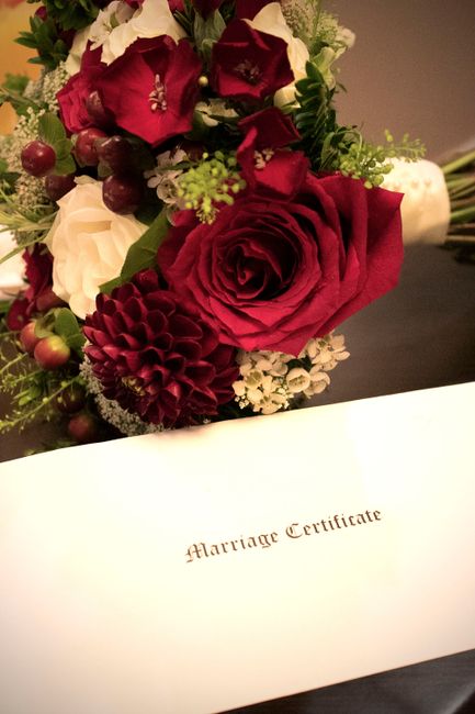 A close-up of a floral arrangement featuring a large, deep red rose at the center, surrounded by burgundy and white flowers including dahlias, small white blooms, and dark red berries. The bouquet is 